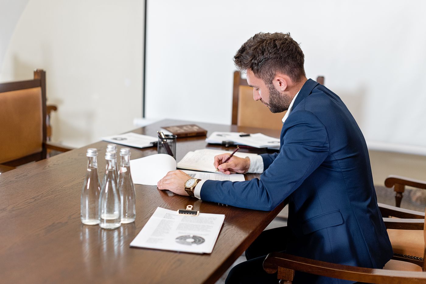Homem de terno azul sentado sozinho a uma mesa comprida de madeira, assinando documentos empilhados diante dele, representando a decisão de formalizar o próprio negócio