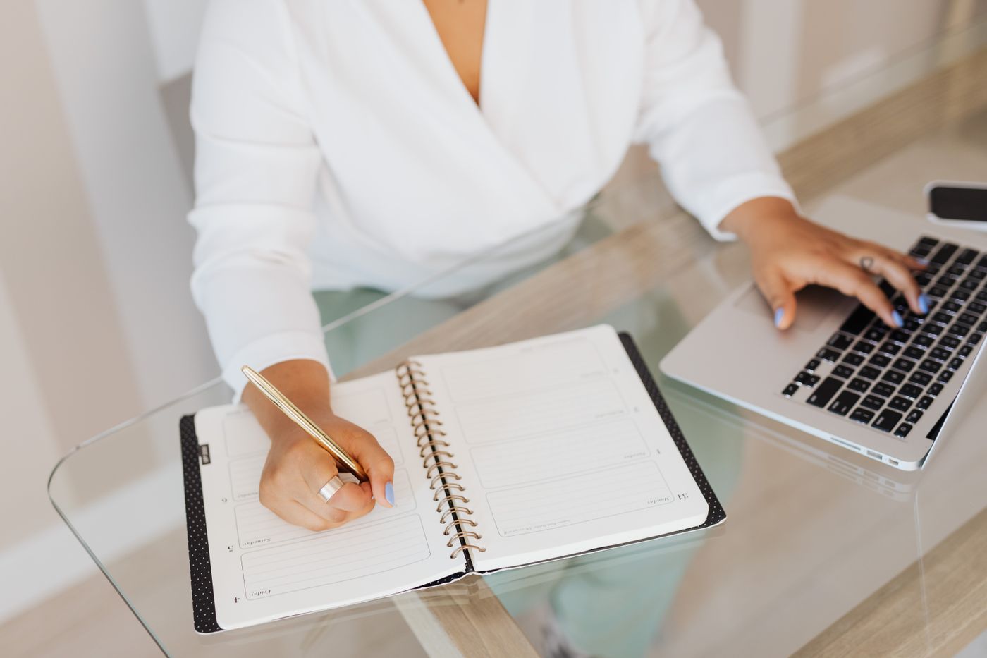 Profissional escrevendo em uma agenda enquanto usa o notebook, planejando o roteiro de estudos para se tornar Educador Financeiro