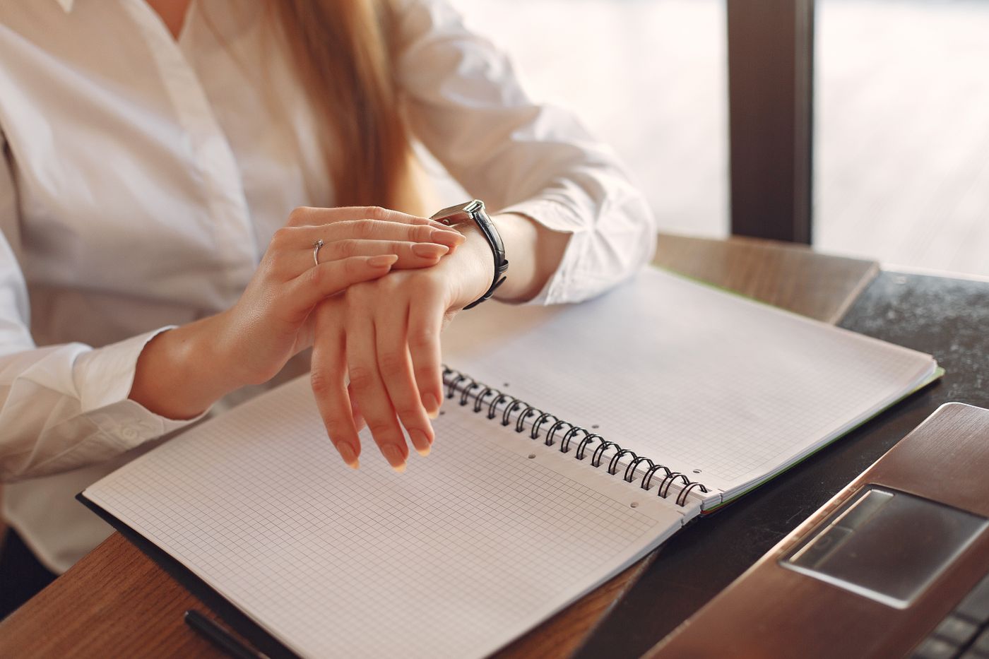 Profissional de camisa branca conferindo o relógio de pulso com um caderno em branco e o notebook sobre a mesa, representando o tempo até se tornar Educador Financeiro