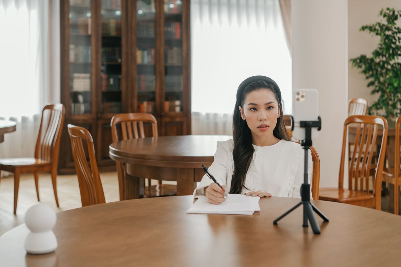Mulher de blusa branca sentada a uma mesa de madeira, escrevendo à mão numa folha enquanto um celular montado num pequeno tripé aponta para ela, com uma estante de livros ao fundo, representando o roteiro escrito antes de ligar a câmera