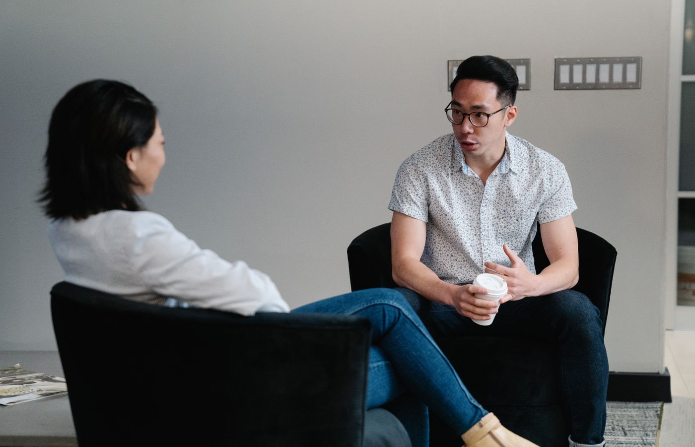Duas pessoas sentadas em poltronas escuras numa conversa atenta, uma delas de costas escutando enquanto a outra fala segurando um copo de café, representando a reunião em que o cliente enfim conta como está a vida financeira dele